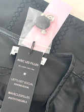 Load image into Gallery viewer, A close-up of a black woven-texture garment with a pink and clear plastic tag attached. The black hangtag reads 'AVEC LES FILLES' and 'LET'S GET SOCIAL / SOYONS SOCIAL' with social handles, and the tag is partially overlaid on a light-colored background.