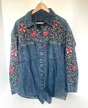 Load image into Gallery viewer, A blue denim button-up shirt jacket hanging on a wooden hanger against a white background. The sleeves and yoke are decorated with a dense multicolor floral print, accented by large pink embroidered flowers.