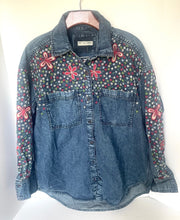 Load image into Gallery viewer, A denim button-up shirt jacket with floral embroidery on the chest and sleeves, hanging on a wooden hanger against a plain white background.