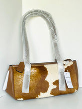Load image into Gallery viewer, A Coach shoulder bag with a brown and cream cowhide pattern, featuring brown leather trim and handles wrapped in protective plastic, with a white brand tag attached.
