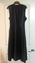 Load image into Gallery viewer, A sleeveless black A-line dress hanging on a wooden hanger against a white paneled door.