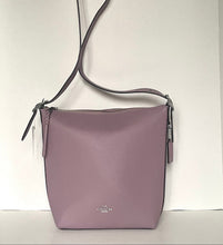 Load image into Gallery viewer, A dusty purple pebbled leather crossbody bag with a matching shoulder strap, hanging against a plain white wall. A small silver-tone Coach logo is visible on the front.