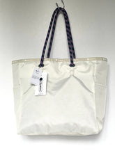 Load image into Gallery viewer, An off-white canvas tote bag with dark patterned shoulder straps, shown hanging against a plain light gray background with its product tag attached.
