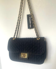 Load image into Gallery viewer, A black quilted Chanel shoulder bag with a gold-tone turn-lock closure and chain strap, hanging against a plain white wall. A black hangtag with the brand name 'CHANEL' is attached.
