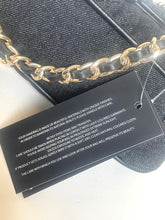 Load image into Gallery viewer, A close-up of a black textured handbag with a gold chain strap, showing a black product care tag with white text.