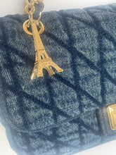 Load image into Gallery viewer, Close-up of a quilted blue denim handbag with a gold-tone Eiffel Tower charm attached near the bottom left, against a plain light background.