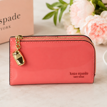 Load image into Gallery viewer, Pink wallet with gold charm on a light surface with flowers and 'kate spade' box in the background.