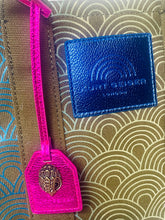Load image into Gallery viewer, Blue leather tag with gold emblem on a textured surface
