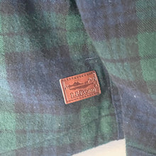 Load image into Gallery viewer, Close-up of a plaid shirt with a leather patch featuring a logo and text.