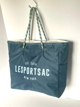 Load image into Gallery viewer, A blue tote bag with green patterned rope handles, featuring the white printed text 'LESPORTSAC EST. 1974 NEW YORK' on the front, hanging from a hook against a plain white wall.