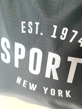Load image into Gallery viewer, Close-up of a dark gray textured fabric with bold white text reading 'EST. 1974 SPORT NEW YORK'.