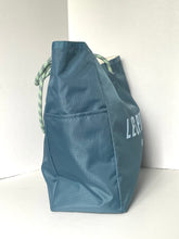 Load image into Gallery viewer, A slate blue reusable tote bag with a drawstring top and white printed text, standing upright against a white background.