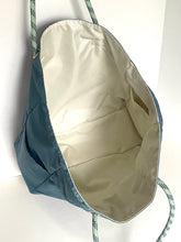 Load image into Gallery viewer, A teal blue nylon drawstring bag with a cream-colored interior lining and green-and-white patterned rope handles, shown against a plain white background.