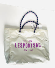 Load image into Gallery viewer, An off-white nylon tote bag with purple text reading 'LESportsac Est. 1974 New York' and purple patterned rope handles, shown on a plain white background with a hangtag attached.