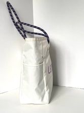 Load image into Gallery viewer, An off-white canvas tote bag with a front pocket, featuring purple embroidered lettering and a dark purple patterned shoulder strap, set against a white corner background.