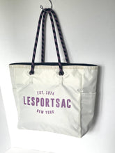 Load image into Gallery viewer, A white canvas tote bag with purple text reading 'EST. 1974 LESPORTSAC NEW YORK' hanging against a plain white wall.