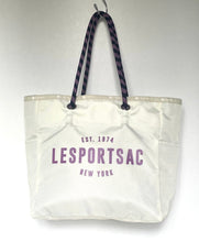 Load image into Gallery viewer, An off-white canvas tote bag with purple text reading 'LESportsac' and 'EST. 1974 NEW YORK' on the front, featuring dark purple patterned shoulder straps, hanging against a plain light gray background.