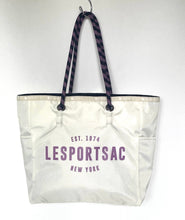 Load image into Gallery viewer, An off-white canvas tote bag with purple text reading 'EST. 1974 LESPORTSAC NEW YORK' hanging from a black-and-white patterned strap on a white wall.