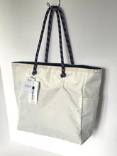 Load image into Gallery viewer, A light beige nylon tote bag with dark purple patterned shoulder straps, hanging from a metal hook against a white wall. The bag has a white hangtag attached to the front.