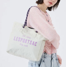 Load image into Gallery viewer, A woman wearing a pink knit sweater and light blue jeans carries a cream canvas tote bag with purple 'LESportsac New York' text and a purple patterned strap against a white background.