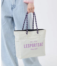 Load image into Gallery viewer, A person wearing light blue jeans holds a cream canvas tote bag with purple 'LESPORTSAC NEW YORK' text and purple patterned shoulder straps against a plain white background.