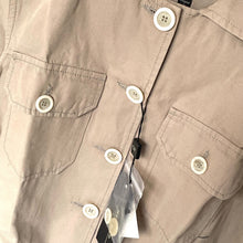 Load image into Gallery viewer, Close-up of a beige shirt with buttons and a tag, showing brand details.
