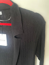 Load image into Gallery viewer, A close-up of a black ribbed knit cardigan sweater hanging on a red hanger against a plain off-white wall. A white hangtag with the brand name 'NAADAM' is visible.