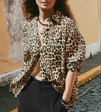 Load image into Gallery viewer, A woman wearing a beige leopard-print button-up shirt and black trousers, standing outdoors against a textured yellow wall.