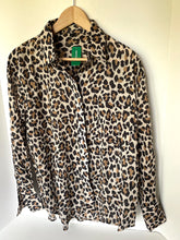 Load image into Gallery viewer, A leopard print long-sleeve button-up shirt hanging on a wooden hanger against a plain off-white wall. The shirt features a beige base with brown and black leopard spots, and a green neck label is visible.