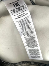 Load image into Gallery viewer, A close-up of a white fabric garment label with black text, including the brand ERL and style information, attached to a light-colored textile with dark stitched trim.