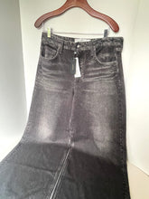 Load image into Gallery viewer, A pair of dark gray washed denim jeans hanging on a wooden hanger against a plain light wall.