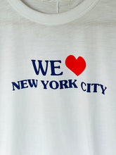 Load image into Gallery viewer, Close-up of a white crewneck t-shirt featuring a graphic print that reads 'WE' followed by a red heart and 'NEW YORK CITY' in navy blue text.