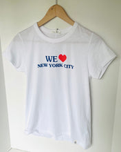 Load image into Gallery viewer, A white short-sleeve crewneck t-shirt hanging on a wooden hanger against a plain white wall. The front of the shirt features a graphic print that reads 'WE' in blue, followed by a red heart, and 'NEW YORK CITY' in blue below.