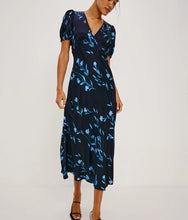 Load image into Gallery viewer, A woman modeling a navy midi dress with a light blue floral print, short puff sleeves, and a wrap-style V-neckline, photographed against a plain light gray studio background.
