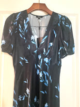 Load image into Gallery viewer, A black short-sleeve wrap dress with a light blue floral print, hanging on a wooden hanger against a cream-colored door.