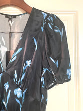 Load image into Gallery viewer, A black sheer blouse with a light blue floral print, featuring puff sleeves and a wrap-style neckline, hanging on a wooden hanger against a cream-colored door.
