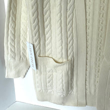 Load image into Gallery viewer, Close-up of a cream-colored cable knit sweater with a brand tag on a white background