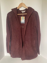Load image into Gallery viewer, A ribbed knit hooded cardigan in a deep burgundy or maroon color, hanging on a wooden hanger against a plain white wall. The cardigan has a single button closure, side pockets, and a hangtag attached to the collar.
