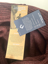 Load image into Gallery viewer, A close-up of a brown knit sweater with two tags attached: a tan paper brand card and a dark gray fabric care label.