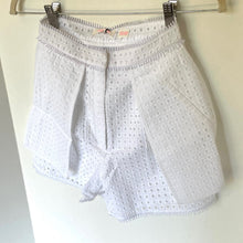 Load image into Gallery viewer, White eyelet shorts with a pleated front and ruffled waistband, hanging from a metal hanger against a plain light background.