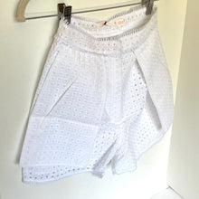 Load image into Gallery viewer, White eyelet shorts with a pleated front, hanging on a hanger against a plain light background.