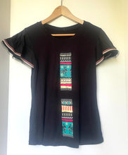 Load image into Gallery viewer, A black short-sleeve t-shirt hanging on a wooden hanger against a plain white wall. The shirt features two vertical panels of colorful geometric and tribal-style patterns in teal, red, yellow, and white on the front, and has striped trim on the sleeves.