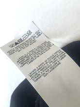Load image into Gallery viewer, A close-up of a white garment care label with black printed text and recycling symbols, resting against a light background with a dark fabric surface below.