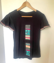 Load image into Gallery viewer, A black short-sleeve t-shirt with a colorful geometric pattern panel on the front and striped frayed trim on the sleeves, hanging on a wooden hanger against a plain white wall.