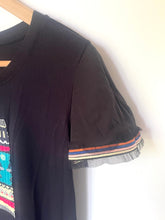 Load image into Gallery viewer, A black short-sleeve t-shirt hanging on a wooden hanger against a plain white wall. The shirt features a colorful geometric pattern on the left chest and a ruffled, sheer mesh trim with orange, blue, and white stripes on the sleeve cuff.