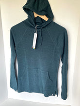 Load image into Gallery viewer, A dark teal long-sleeve hooded top hanging on a wooden hanger against a plain white wall, with a white hangtag attached near the neckline.
