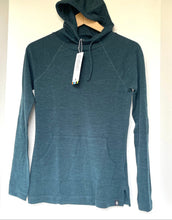 Load image into Gallery viewer, A dark teal heathered long-sleeve hooded top hanging on a wooden hanger against a plain off-white wall. The garment has drawstrings, raglan sleeves, a kangaroo pocket, and a hangtag attached near the collar.