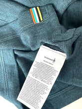 Load image into Gallery viewer, Close-up of a teal knit garment with a white Smartwool care tag attached near the collar, showing a small striped fabric label above it.