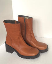 Load image into Gallery viewer, A pair of tan leather ankle boots with black soles and block heels, shown on a light gray surface against a white background.