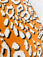 Load image into Gallery viewer, A close-up of a leopard print fabric featuring a bold orange background with irregular white spots outlined in black.